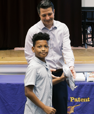  A student looks towards the camera smiling as a man standing behind him hands him a piece of paper.